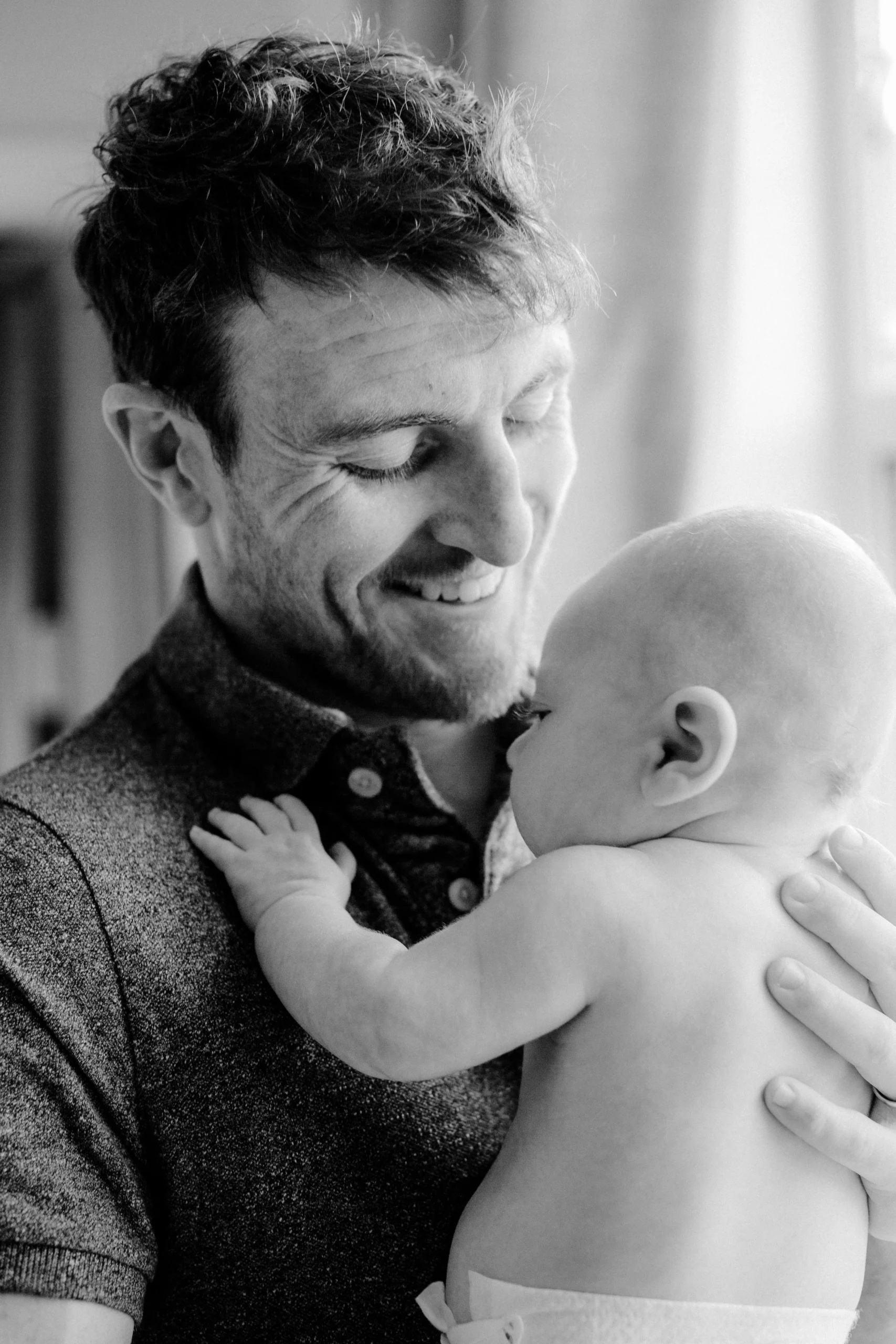 A black-and-white photo of a man smiling and holding a baby close to his chest. The baby is shirtless and touching the man's face, with both looking at each other lovingly.