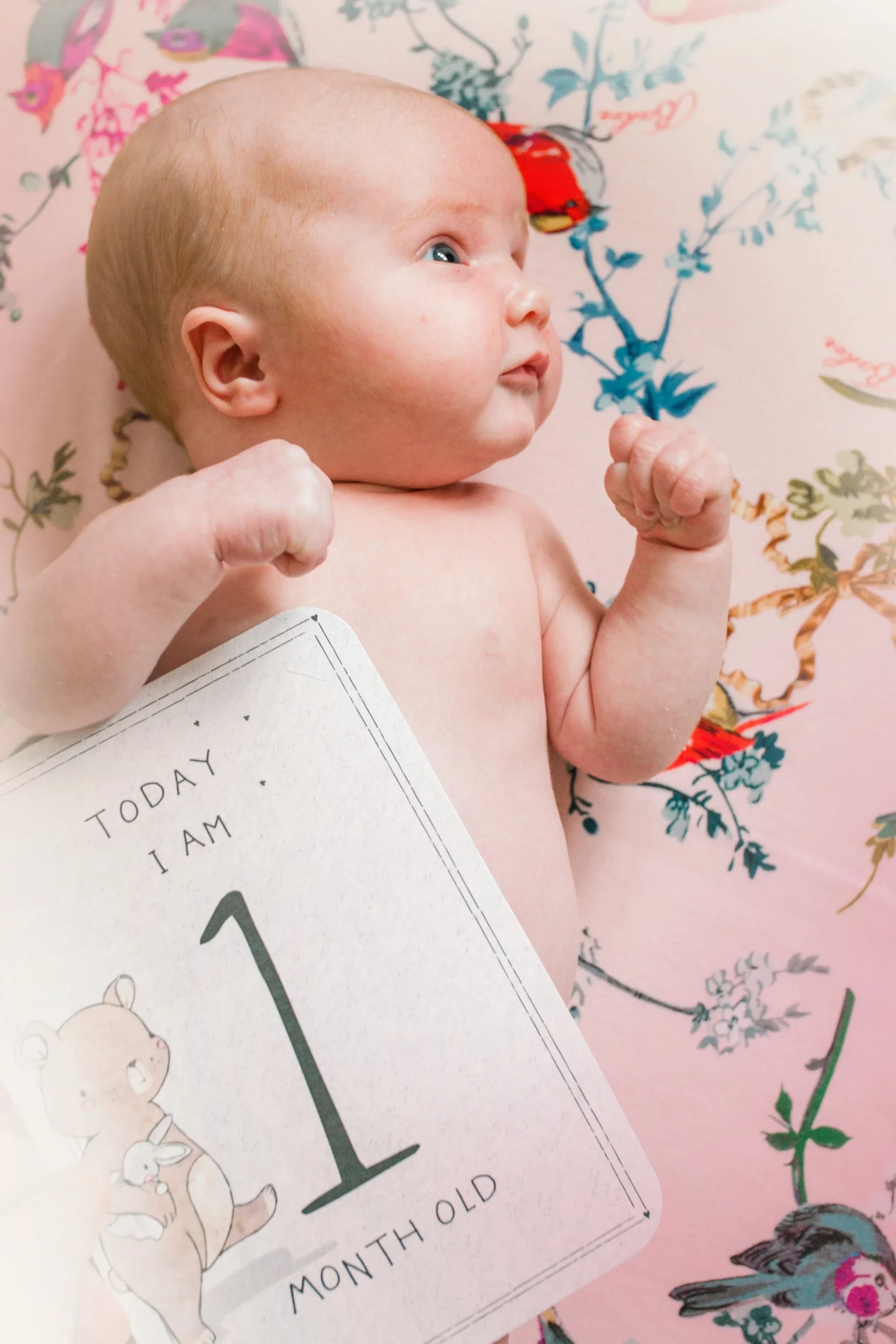A young baby lying on a floral blanket, showing their chest and arms, with fists clenched in a cute pose, beside a sign indicating the baby is 1 month old.