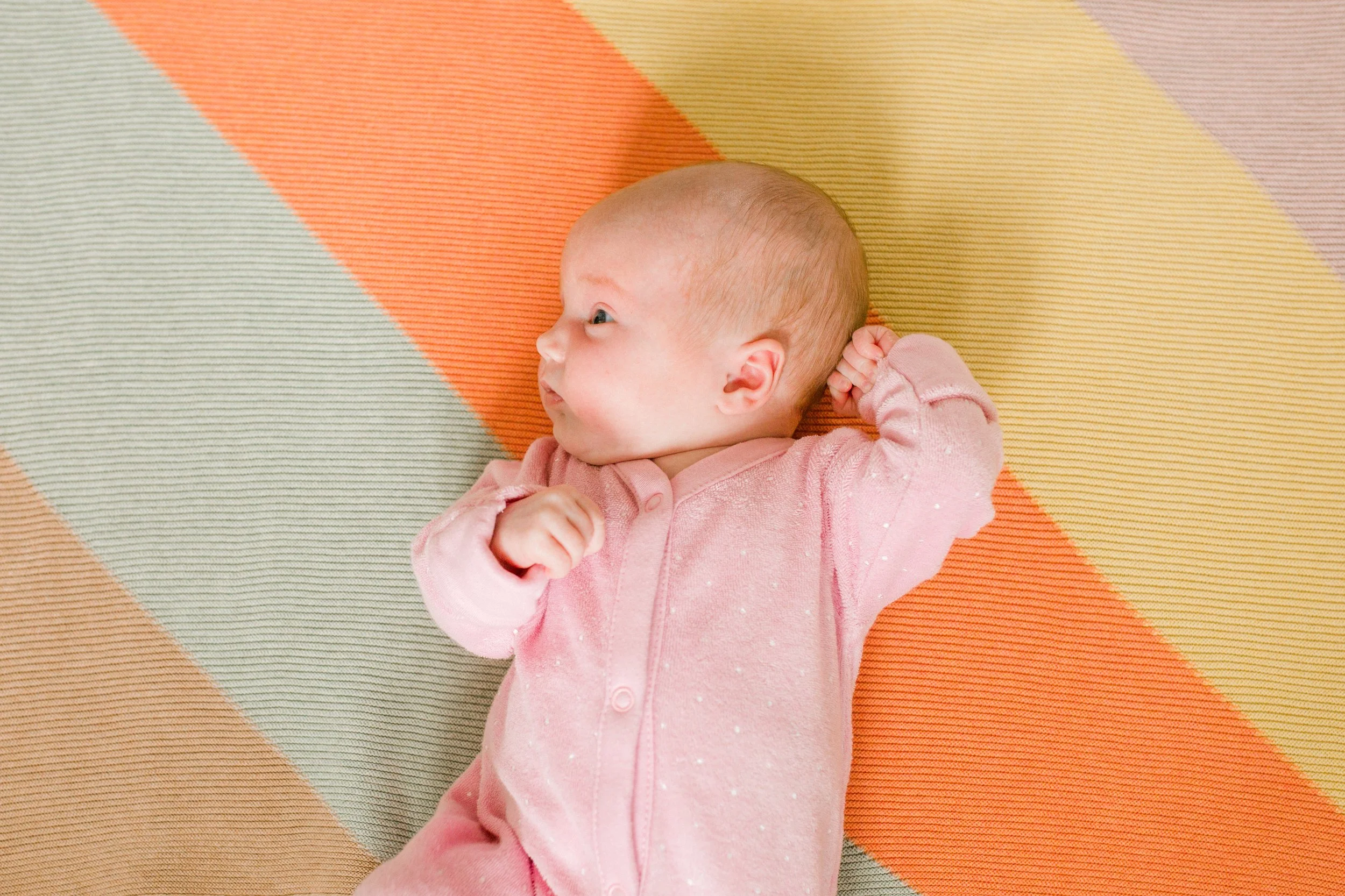 Baby lying on colorful striped mat, wearing a pink outfit, looking to the side with a curious expression.