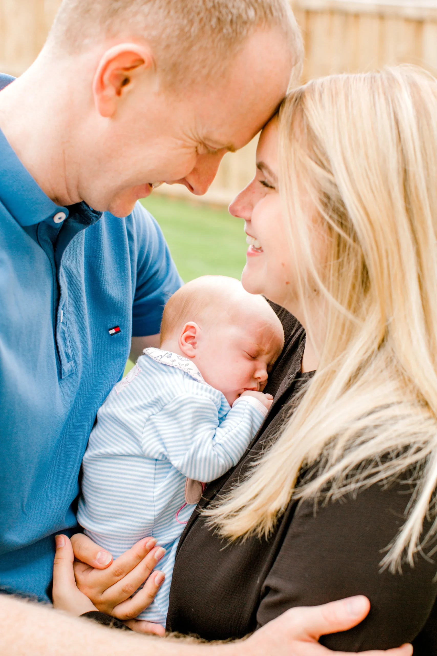A couple is smiling and touching foreheads with their baby, who is sleeping peacefully in the woman's arms.