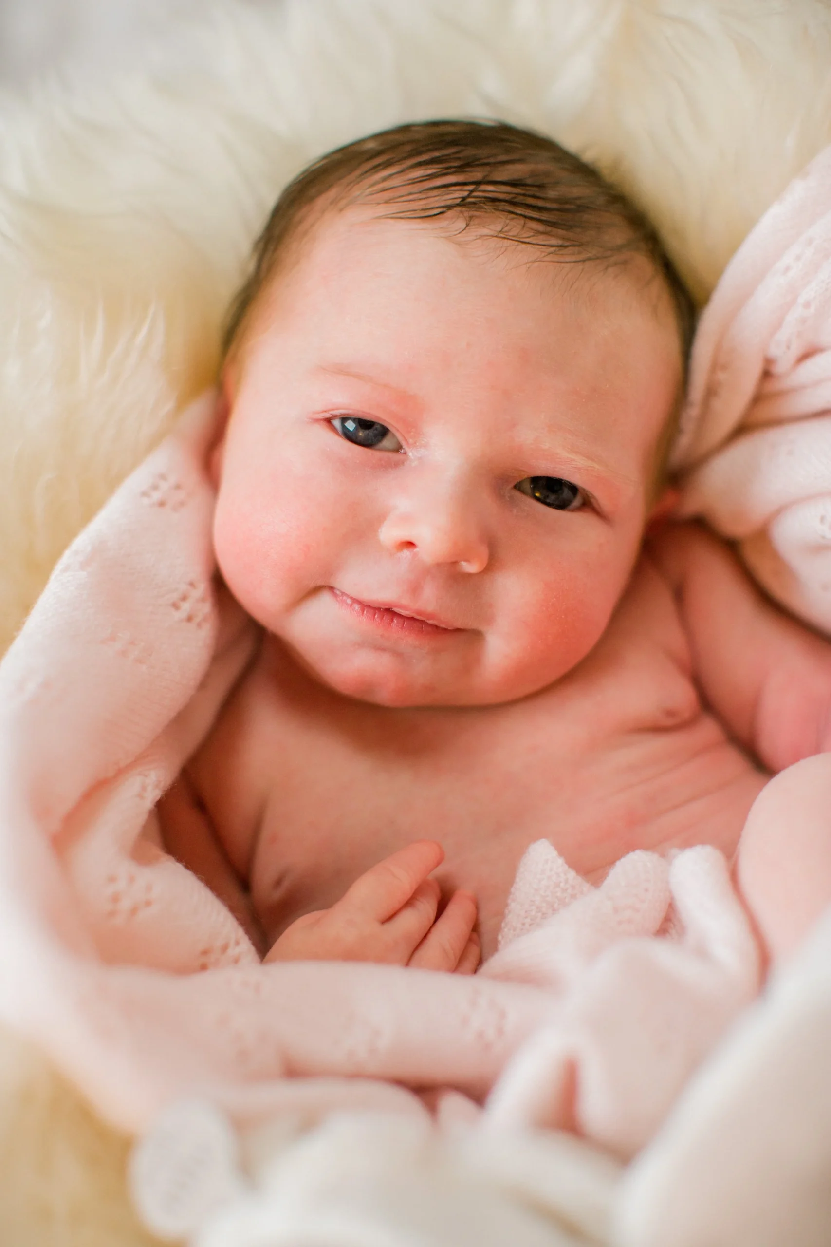 Close-up of a smiling newborn baby with dark hair, lying on a soft cream-colored surface, wrapped in a pink blanket.