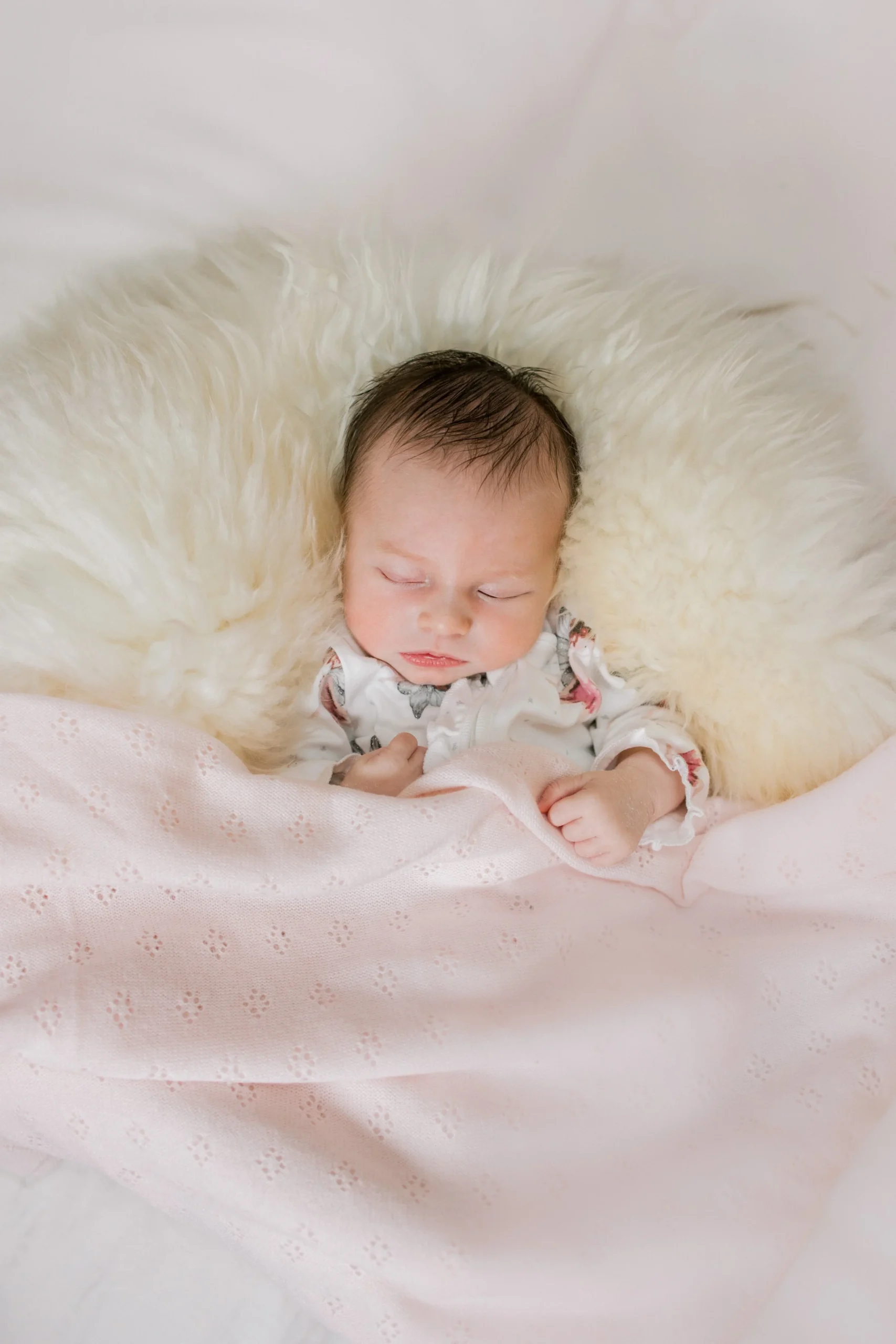 A sleeping baby with wet hair lying on a fluffy white blanket, partially covered with a pink blanket.