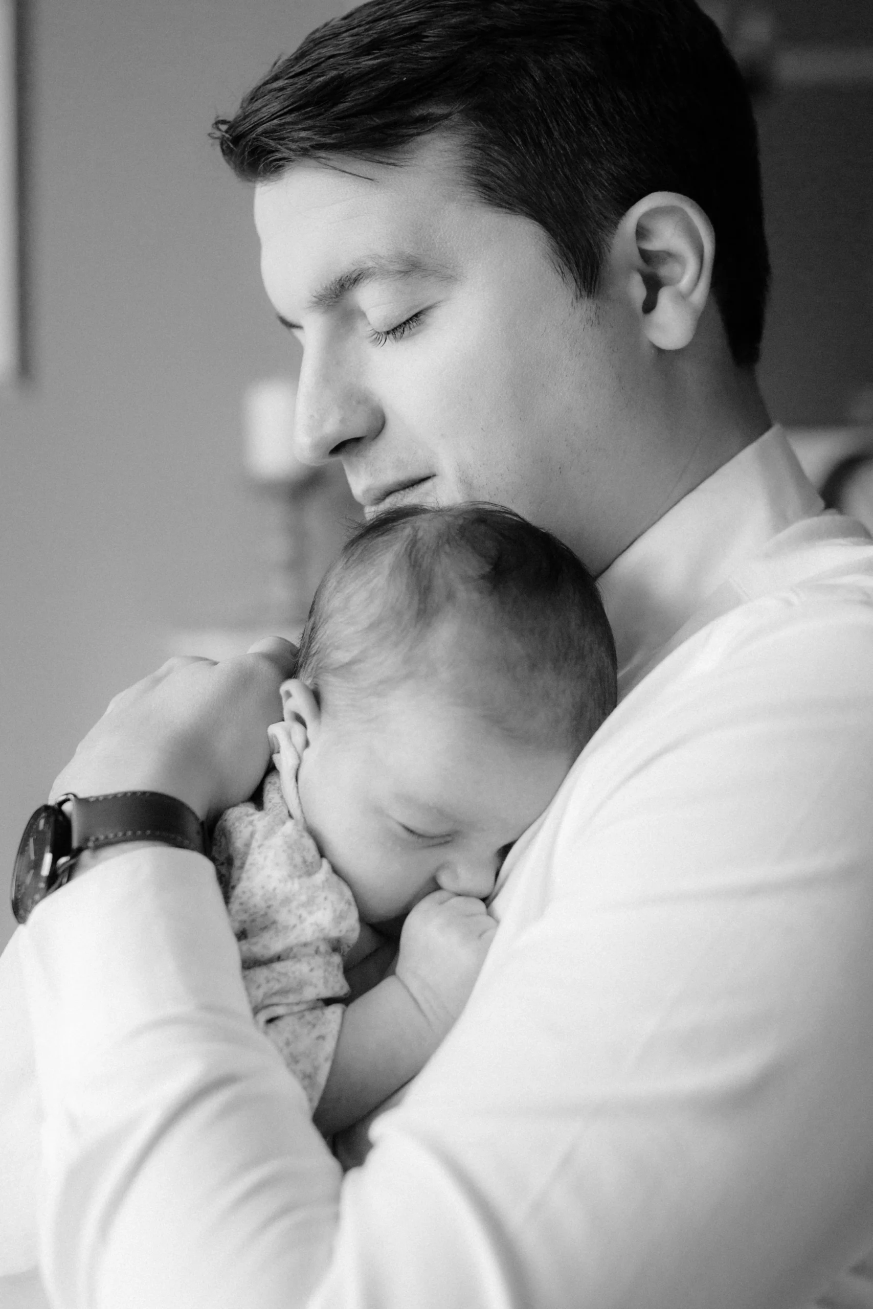 A black and white photo of a young man holding a sleeping baby close to his chest, with his eyes closed and a peaceful expression.