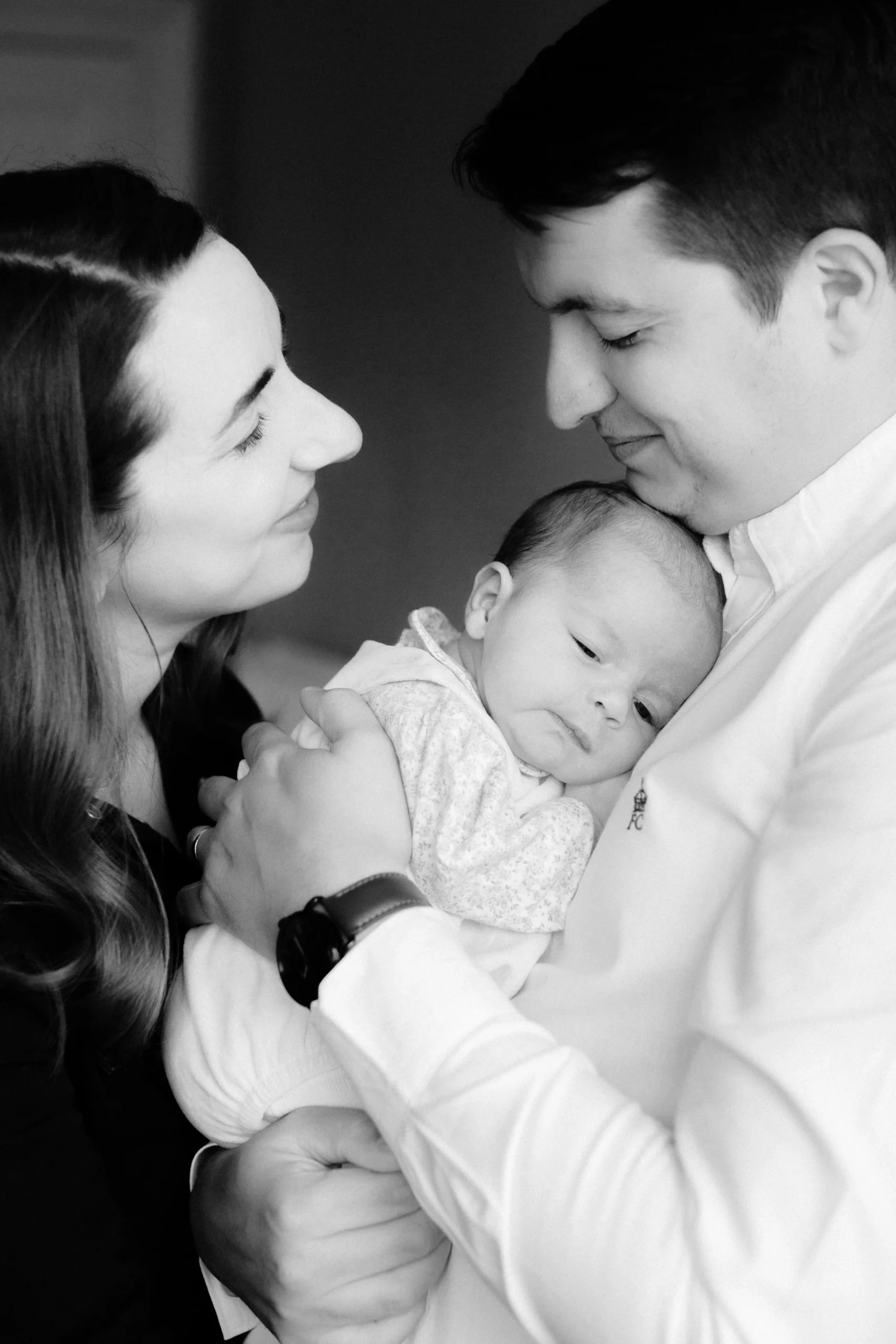 A family moment showing a woman, man, and baby, all with close heads together, with the woman and man smiling and the baby looking thoughtful.