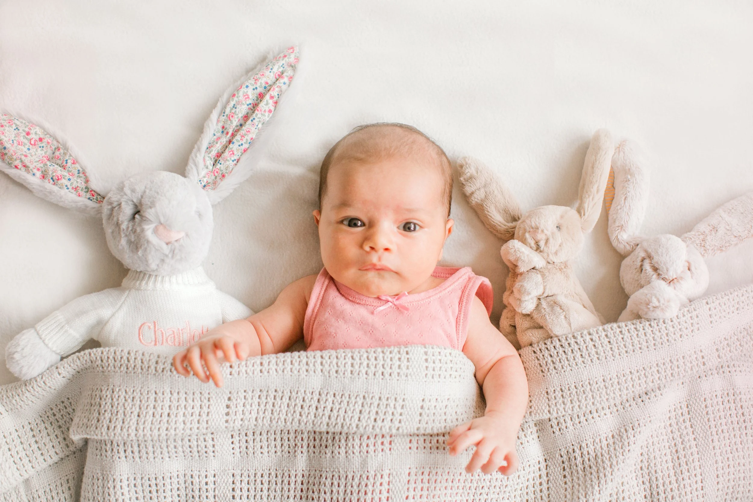 A baby lying on a bed with two plush bunny stuffed animals, one on each side, and a white blanket partially covering the baby.