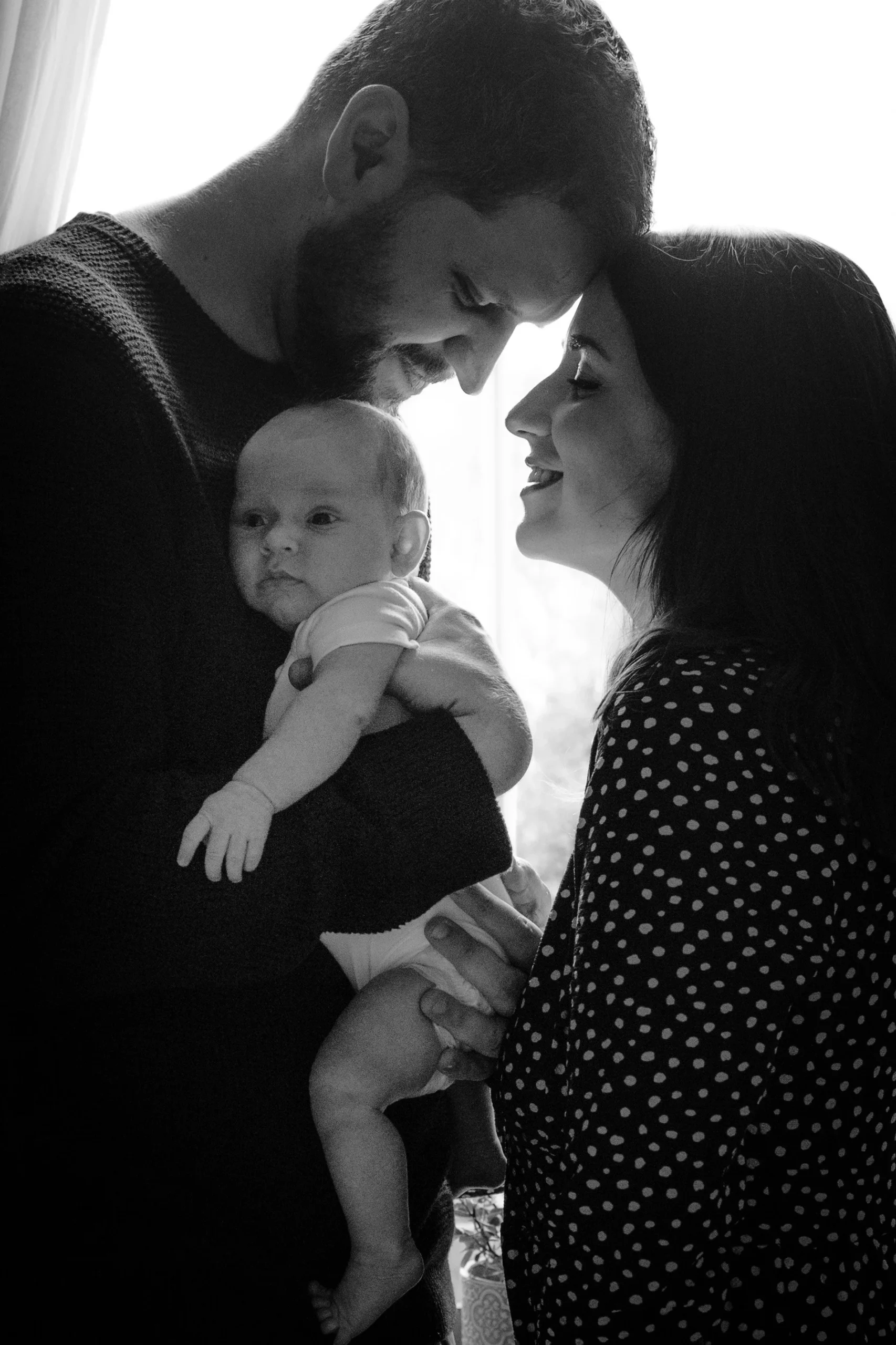 A black and white photo of a happy family with a man and woman seeing each other with their foreheads touching, holding a baby together near a window.