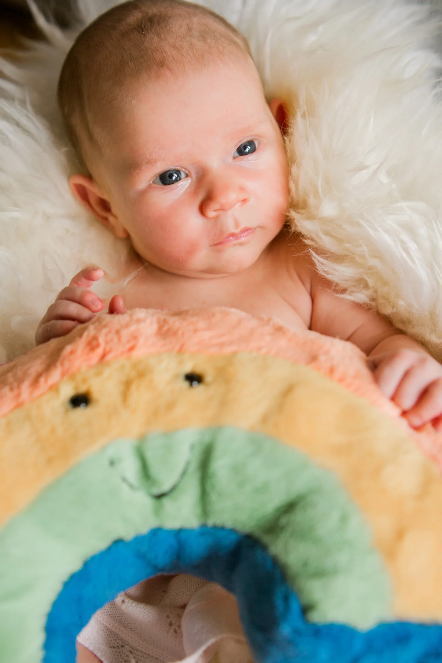 A baby lying on a white fluffy surface, holding a colorful blanket with a smiley face. The baby has blue eyes and light brown hair.