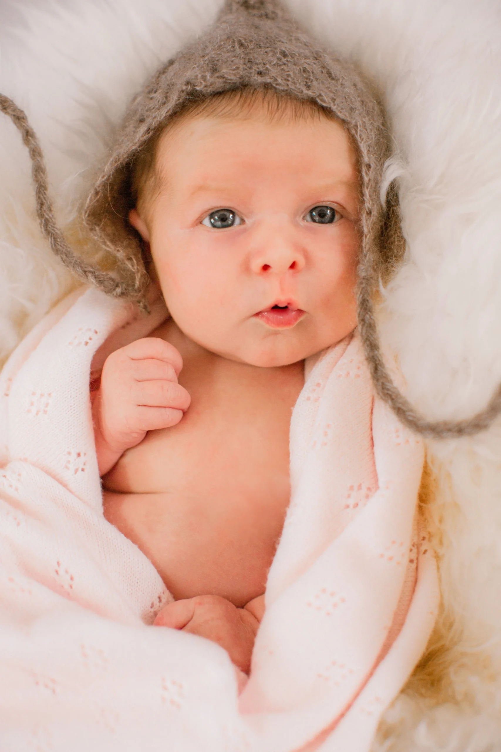 A baby with blue eyes, wearing a brown wool hat with tassels, wrapped in a white knitted blanket with small eyelet patterns, lying on a fluffy cream-colored surface.