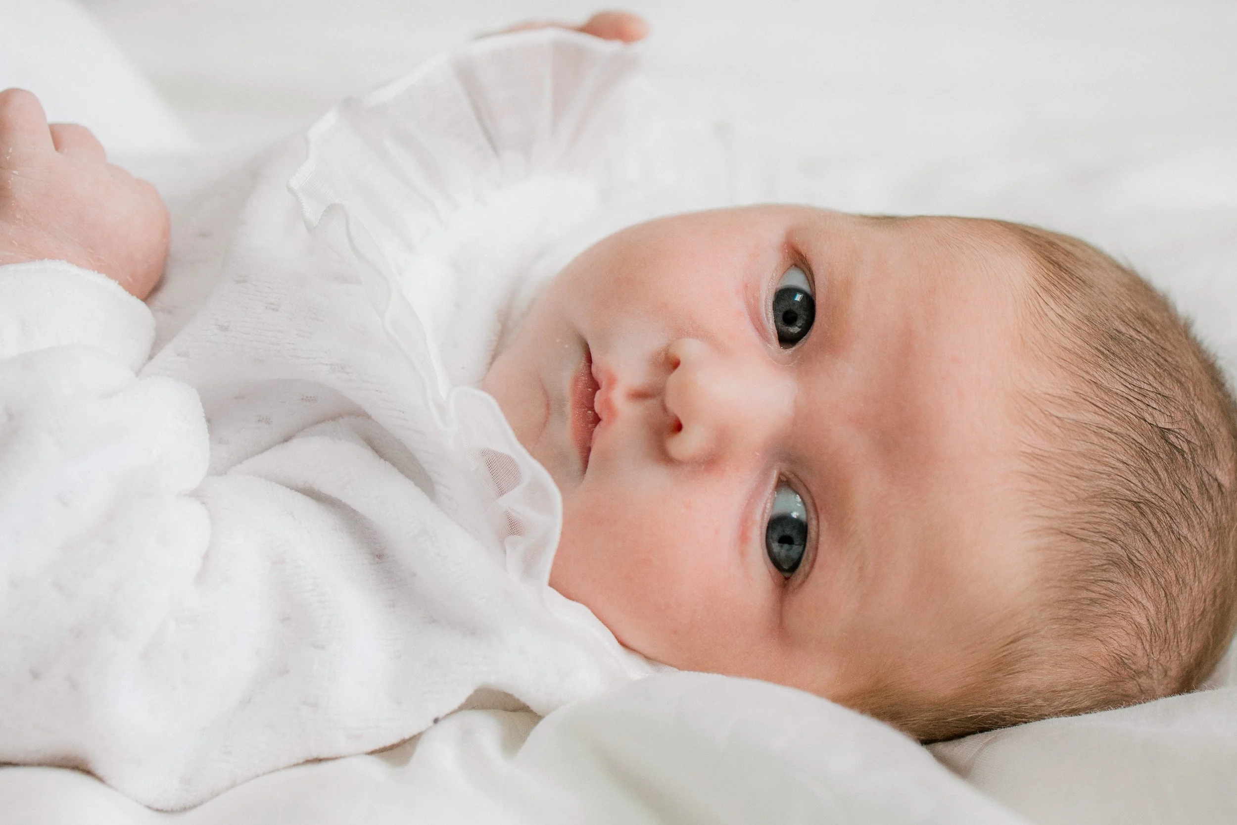 Close-up of a baby lying on a white surface, wearing a white outfit with ruffled collar, looking off to the side.
