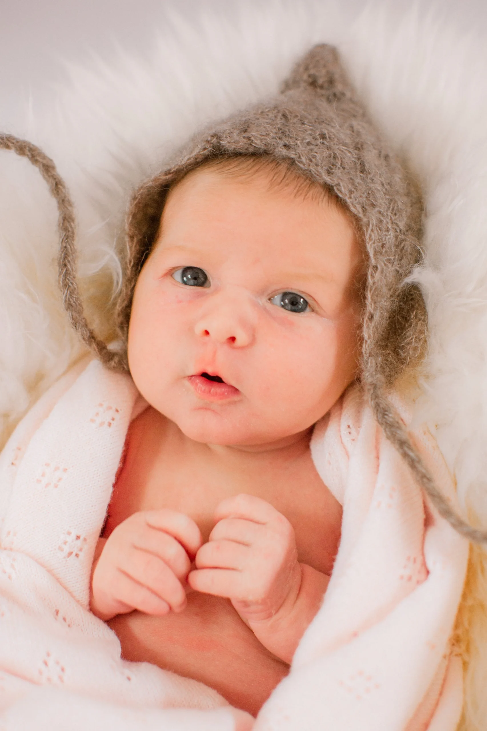 A baby with blue eyes, wearing a fuzzy brown hat with ear flaps and string ties, wrapped in a soft pink blanket, lying on a fluffy white surface.