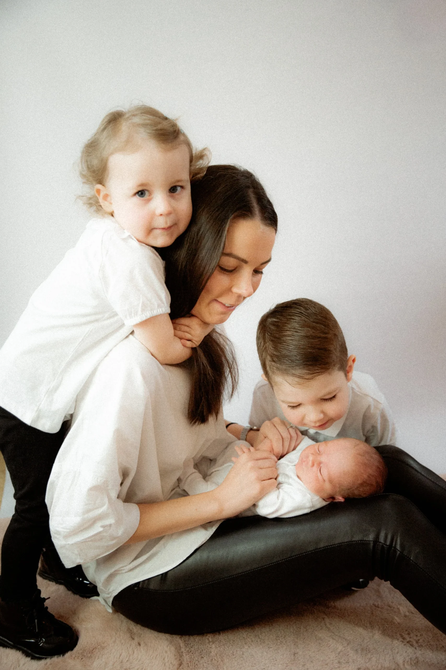 A woman with two children and a newborn baby, all dressed in light-colored clothing, gathered around on a black chair. The woman holds the baby and the children look and touch the baby affectionately.