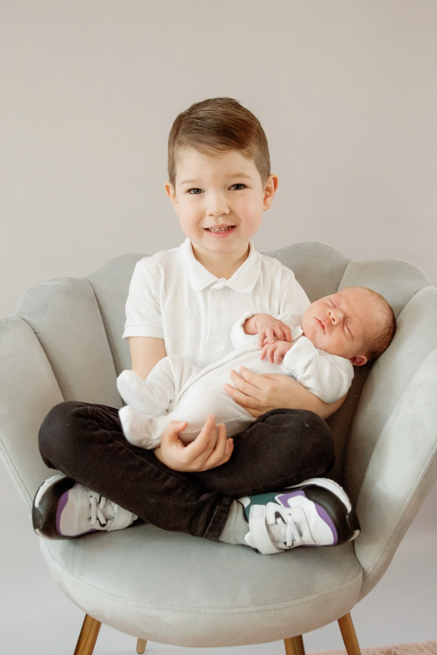 A young boy with brown hair sitting cross-legged on a modern beige chair, holding a newborn baby in his arms. The baby is dressed in a white onesie and appears to be sleeping.