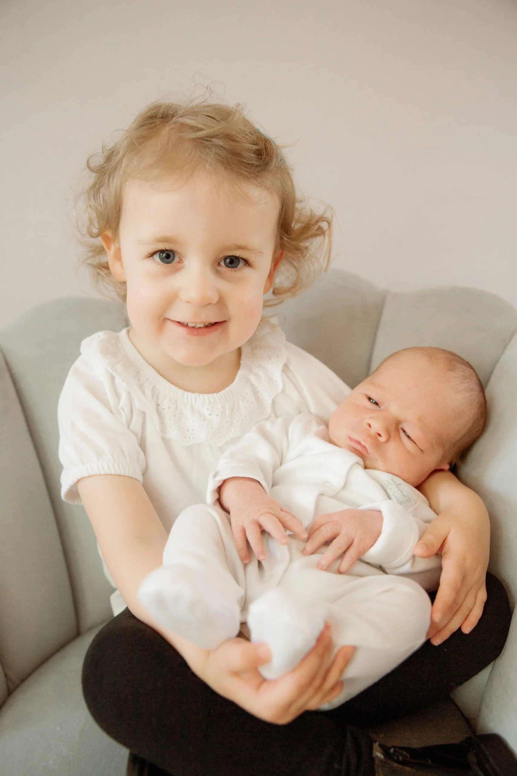 A young girl with curly blonde hair and blue eyes smiling and holding a newborn baby with light skin and short hair, seated on a light-colored chair against a white wall.