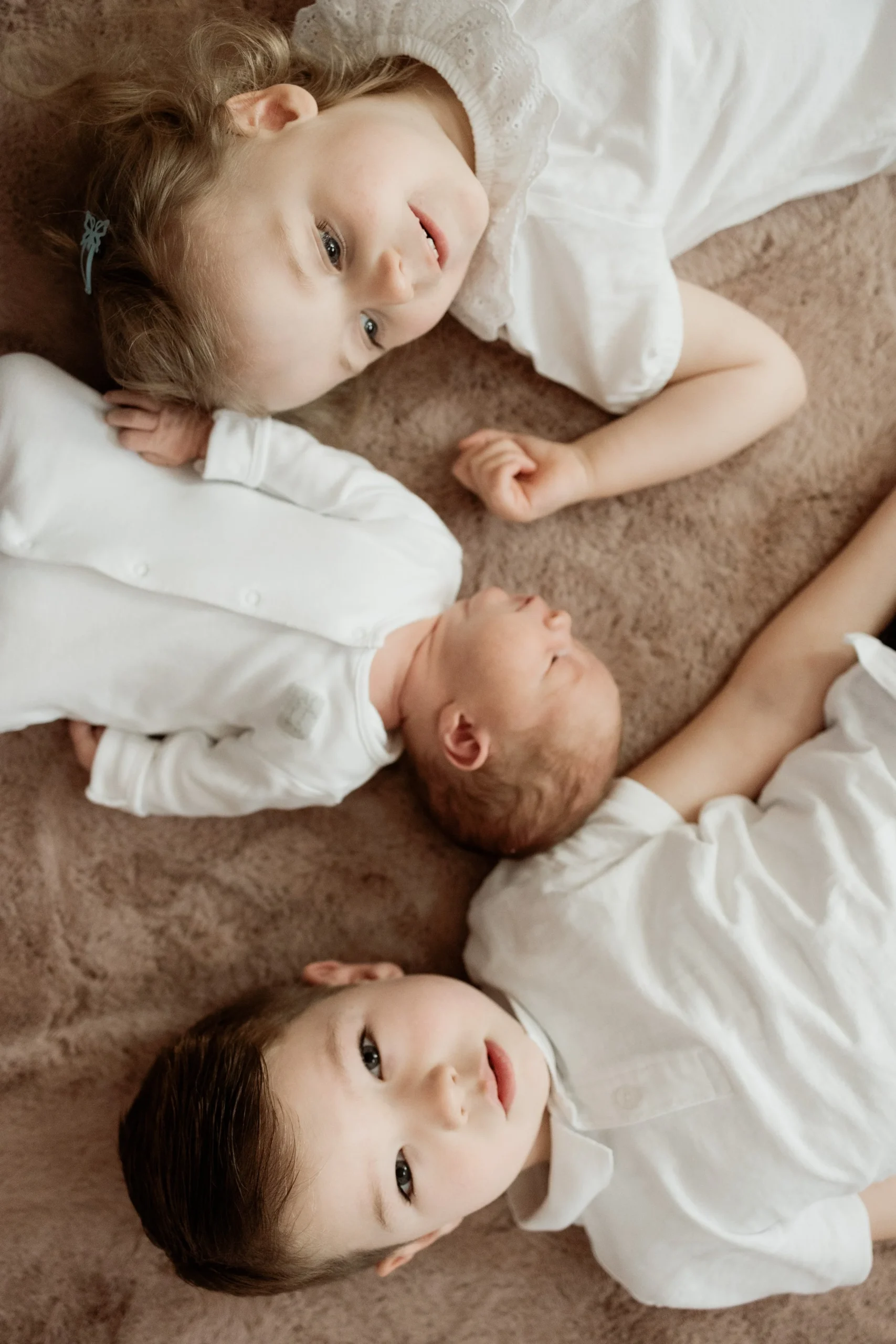 Three young children lying on a soft brown carpet, one girl and one boy with a baby, all dressed in white, resting and looking at the camera.