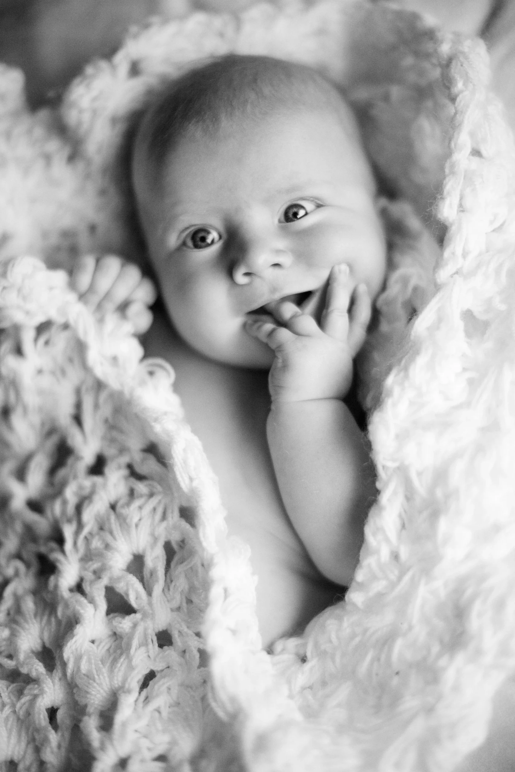 A smiling baby lying on a knitted blanket, looking at the camera with one hand near their mouth.