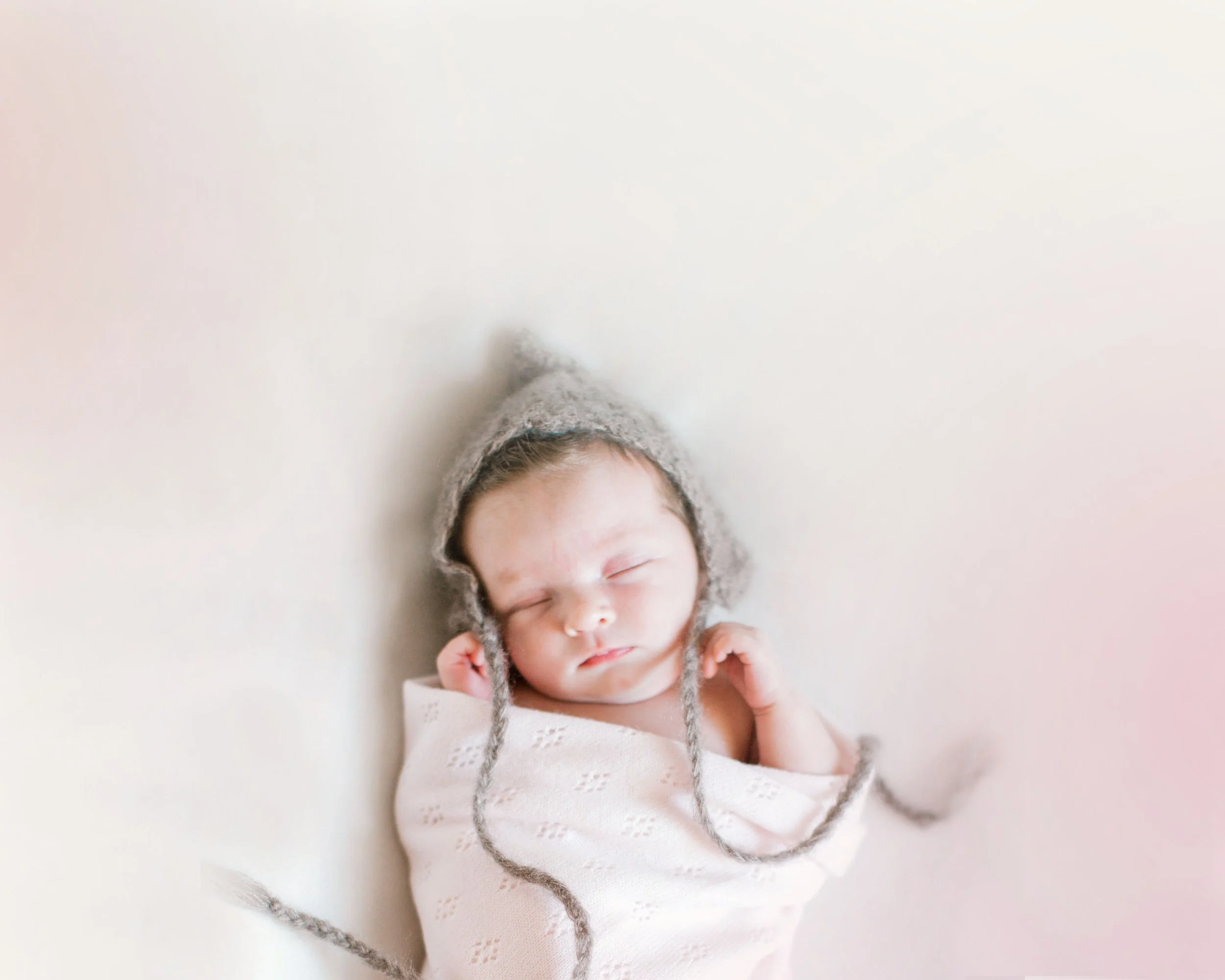 A sleeping newborn baby swaddled in a white blanket, wearing a gray knitted hat with braided strings.