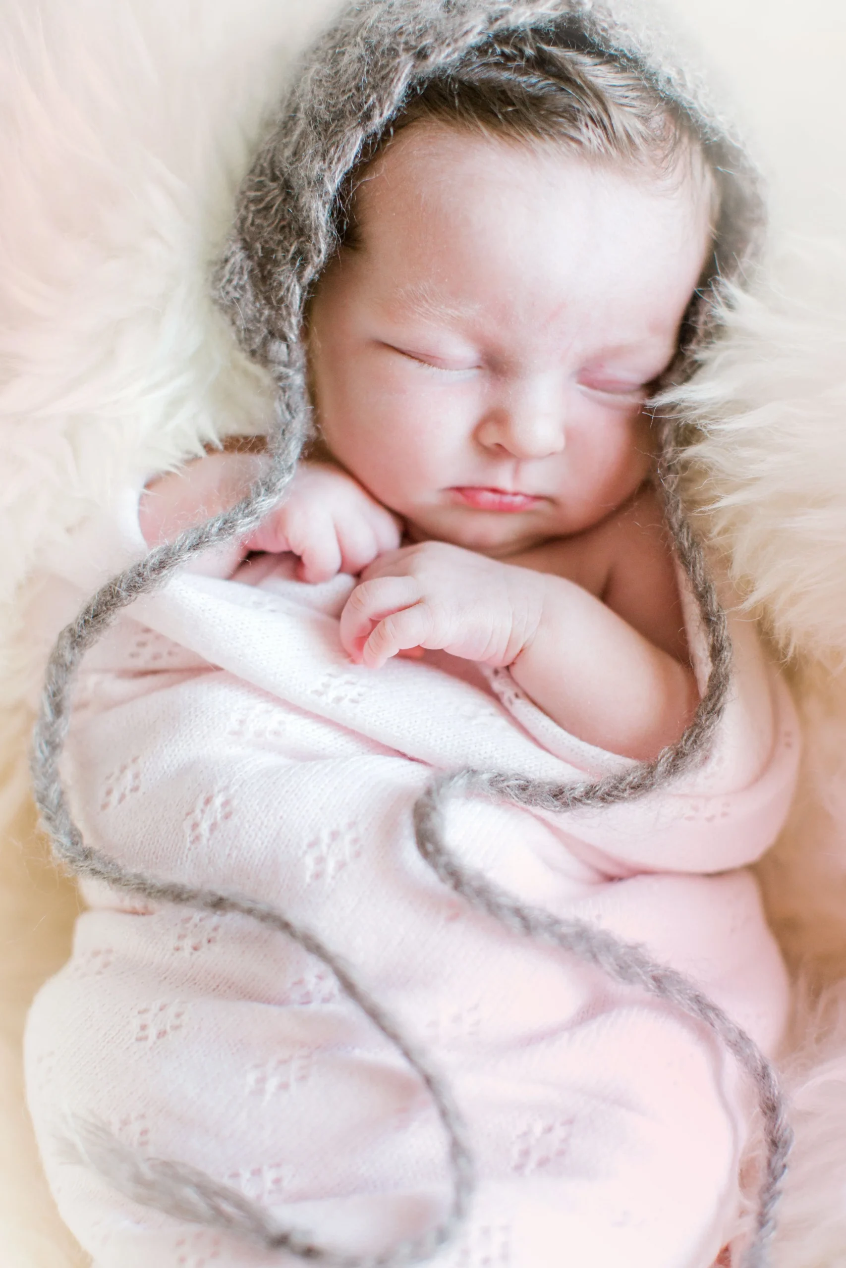 A sleeping baby wrapped in a pink blanket, wearing a cozy knitted hat with long braids, lying on a soft, fluffy surface.